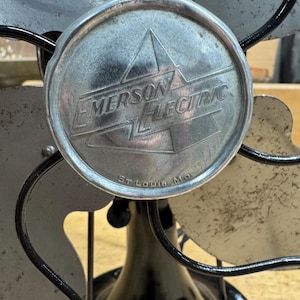May include: Vintage Emerson Electric fan with a black base and cage, and silver fan blades. The center of the fan features a circular emblem with the Emerson Electric logo and the text "St. Louis Mo."