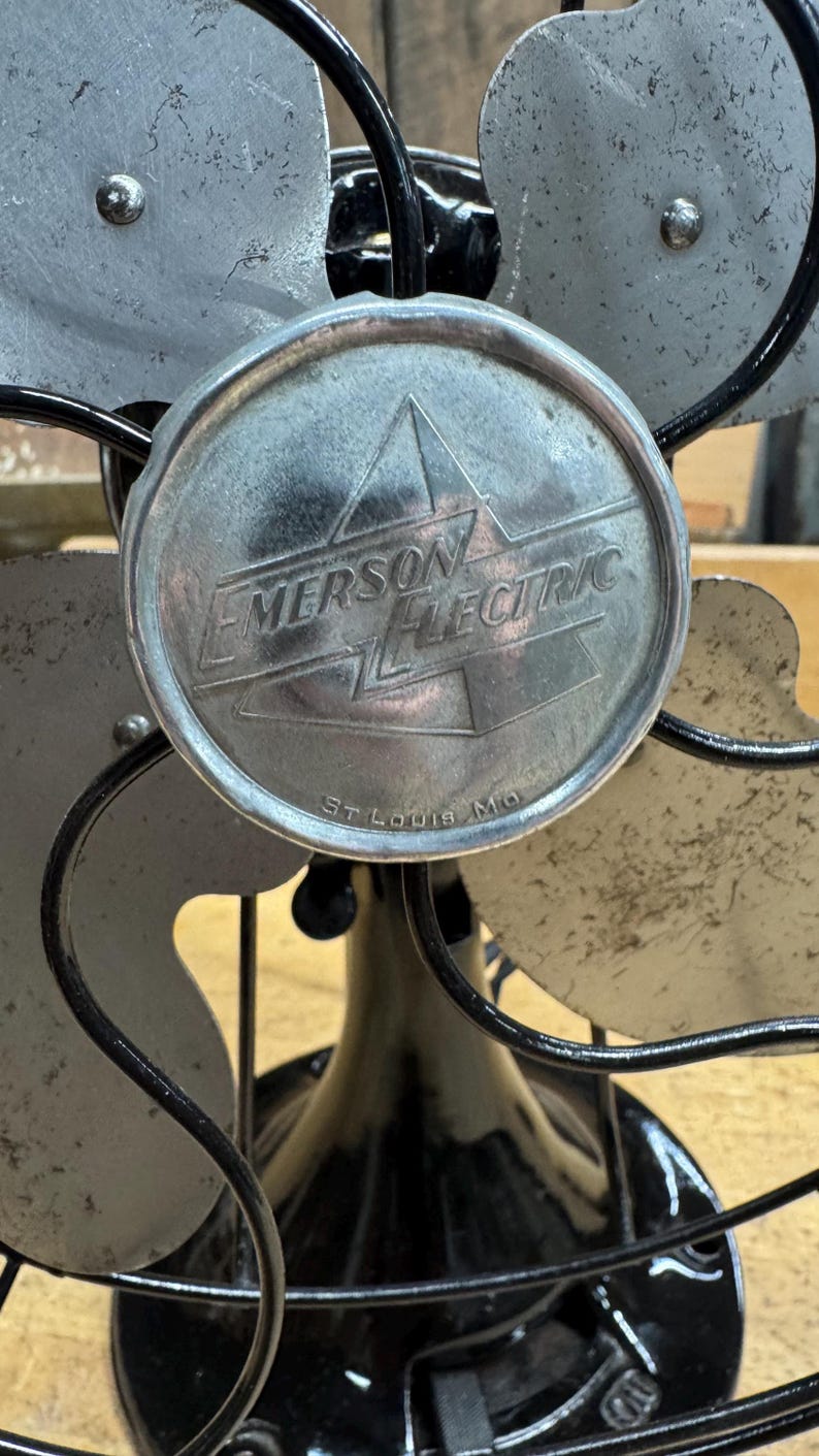 May include: Vintage Emerson Electric fan with a black base and cage, and silver fan blades. The center of the fan features a circular emblem with the Emerson Electric logo and the text "St. Louis Mo."