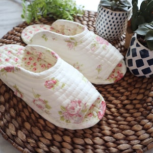 May include: A pair of white quilted slippers with a floral pattern. The slippers are on a woven brown mat.