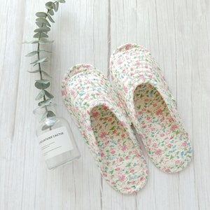 May include: A pair of white floral print slippers with pink and blue flowers. The slippers are on a white wooden surface.