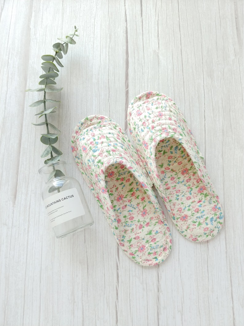 May include: A pair of white floral print slippers with pink and blue flowers. The slippers are on a white wooden surface.