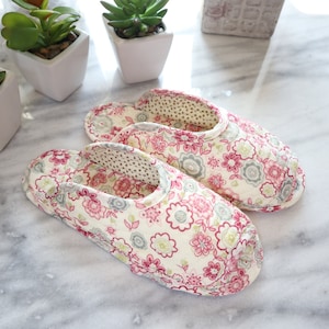 Può includere: Un paio di pantofole con stampa floreale su sfondo crema e fiori rosa e verdi. Le pantofole sono realizzate in tessuto morbido e offrono una calzata confortevole.