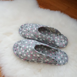 May include: A pair of gray and white polka dot slippers with pink accents. The slippers are on a white fluffy surface.