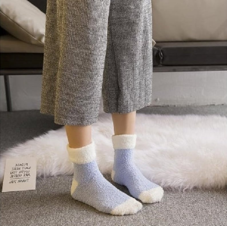 May include: A pair of fuzzy socks with a white cuff and light blue fuzzy fabric. The socks are worn by a person wearing grey pants.
