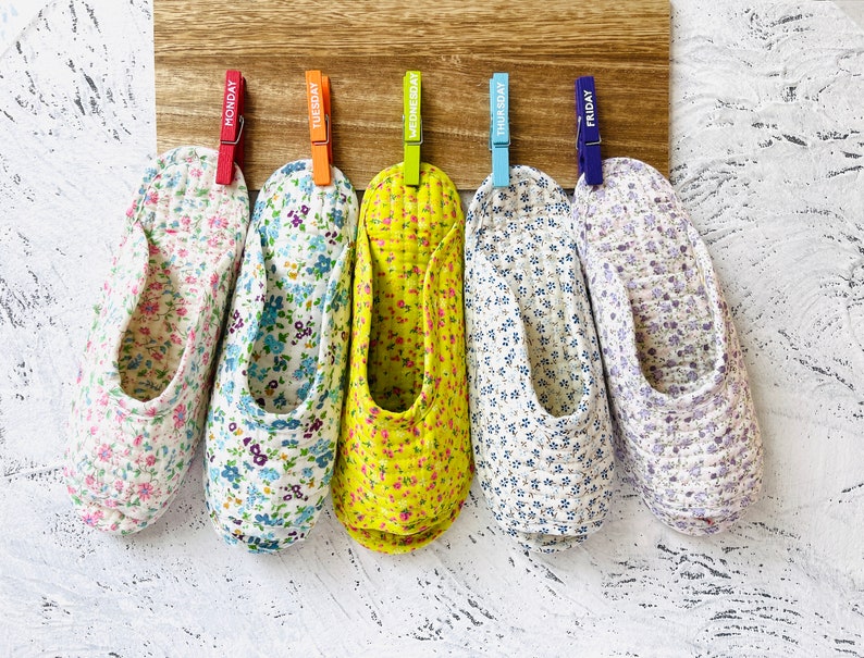 May include: Five pairs of floral patterned slippers hanging on a wooden board with clothespins labeled Monday through Friday.
