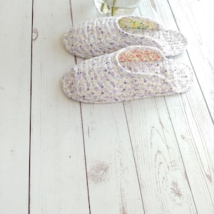May include: A pair of white floral print slippers with purple flowers. The slippers are on a white wooden surface.