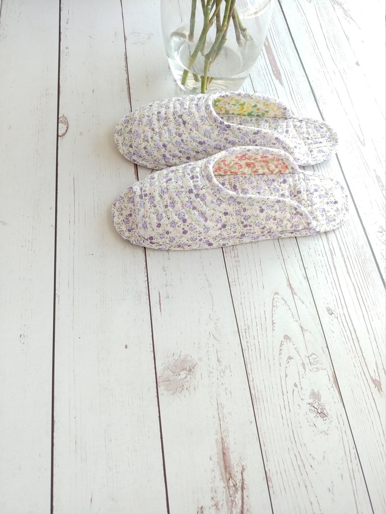 May include: A pair of white floral print slippers with purple flowers. The slippers are on a white wooden surface.