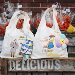 Könnte beinhalten: Zwei weiße Tragetaschen mit Illustrationen und Text. Eine Tasche zeigt eine Frau in einem Kleid, den Text "SOZAKKA Spice latte" und einen Schlüsselanhänger in Form eines Milchkartons. Die andere Tasche hat eine Person auf einem Fahrrad, den Text "SEASON of SOZAKKA" und einen Blumen-Schlüsselanhänger.