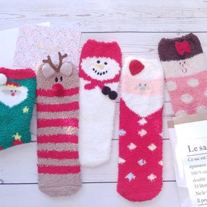 Könnte beinhalten: Eine Auswahl an flauschigen Weihnachtssocken. Designs umfassen den Weihnachtsmann, ein Rentier, einen Schneemann und eine rosa Socke mit Punkten. Die Socken liegen auf einer hellen Holzoberfläche.