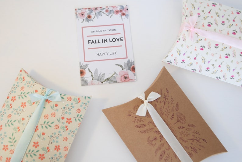 May include: Three pillow boxes with different floral patterns and ribbons. The boxes are white with pink flowers, beige with orange flowers, and brown with a brown floral design. The boxes are tied with ribbons in different colors. The boxes are perfect for packaging small gifts or favors.
