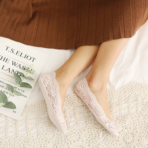Könnte beinhalten: Ein Paar hellrosa, spitzenbesetzte Füßlinge. Die Socken liegen auf einer weißen Oberfläche mit einem Buch mit dem Titel "The Waste Land" von T.S. Eliot. Die Socken haben ein zartes florales Spitzendesign.