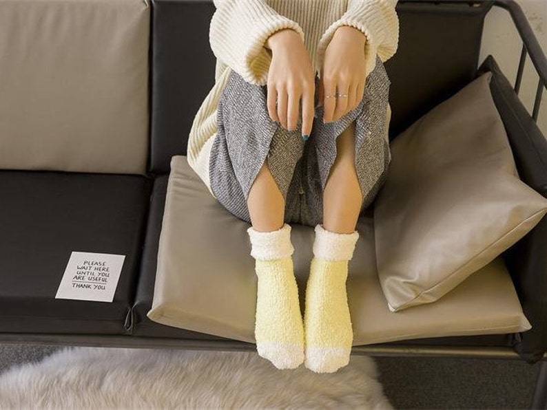 May include: A woman wearing a white sweater and gray pants sits on a couch with a sign that reads "Please wait here until you are called. Thank you." She is wearing yellow fuzzy socks.