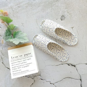 May include: A pair of white and blue floral print slippers on a white textured background. The slippers are open-toed and have a simple design. A small brown and white notepad with text in French is in the lower left corner of the image.