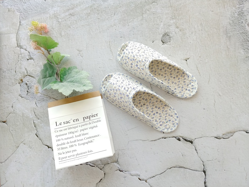 May include: A pair of white and blue floral print slippers on a white textured background. The slippers are open-toed and have a simple design. A small brown and white notepad with text in French is in the lower left corner of the image.