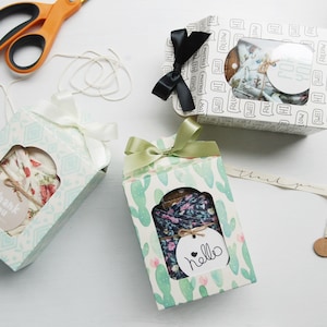 May include: Three gift boxes with different patterns and colors. The boxes are tied with ribbon and have a small window to see the contents. The boxes are on a white surface with spools of twine and ribbon.