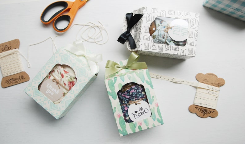 May include: Three gift boxes with different patterns and colors. The boxes are tied with ribbon and have a small window to see the contents. The boxes are on a white surface with spools of twine and ribbon.