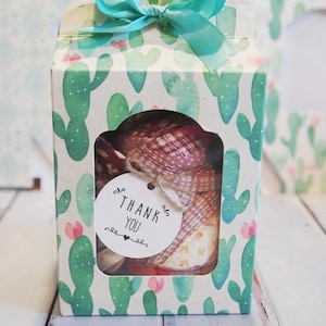 May include: A small gift box with a clear window, featuring a cactus print and a turquoise ribbon. The box contains a stack of fabric squares with a variety of patterns. A small tag with the text "THANK YOU" is attached to the fabric.