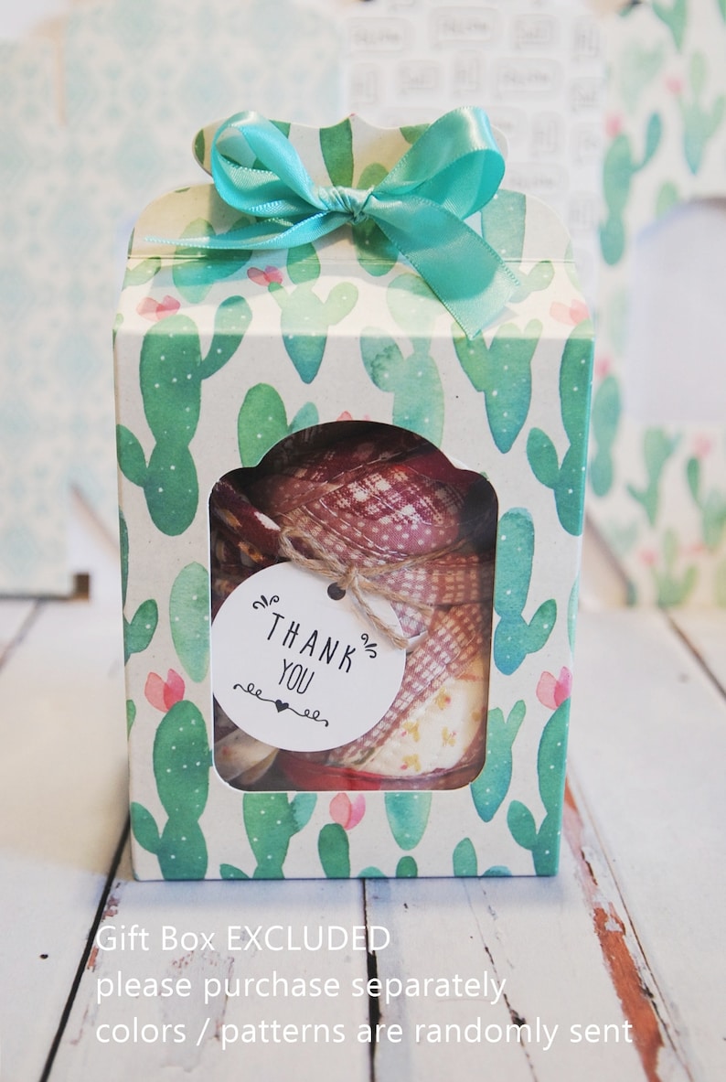 May include: A small gift box with a clear window, featuring a cactus print and a turquoise ribbon. The box contains a stack of fabric squares with a variety of patterns. A small tag with the text "THANK YOU" is attached to the fabric.