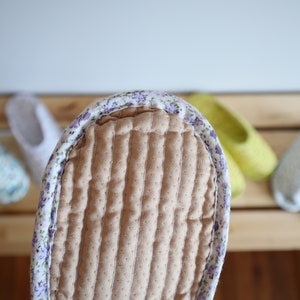 May include: Close-up of a brown quilted slipper with a floral print border. The slipper is on a wooden surface with other slippers in the background.