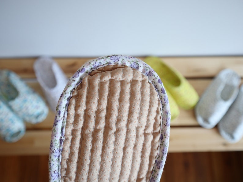 May include: Close-up of a brown quilted slipper with a floral print border. The slipper is on a wooden surface with other slippers in the background.