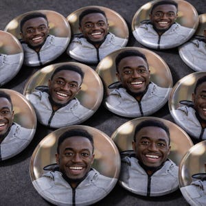 Peut inclure: Plusieurs badges ronds repr&eacute;sentant une personne souriante portant un sweat &agrave; capuche gris. Les badges sont dispos&eacute;s sur une surface sombre, le visage et le haut du corps de la personne &eacute;tant visibles sur chaque badge.