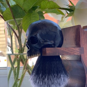 May include: A black skull-shaped shaving brush handle with a black and white badger hair brush. The handle is mounted on a wooden stand.