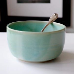 May include: A light teal ceramic bowl with a silver spoon inside. The bowl has a rounded shape and a smooth, glossy finish. The spoon has a decorative handle. The bowl is sitting on a white surface.