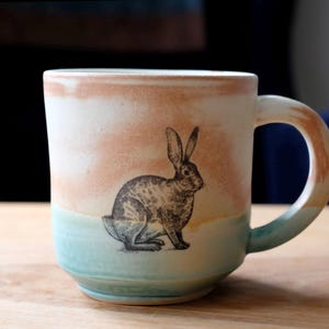 May include: A ceramic mug with a rabbit illustration. The mug features a gradient design with colors of peach, white, and teal. The rabbit is depicted in a detailed, vintage style. The handle is also painted with the same gradient.
