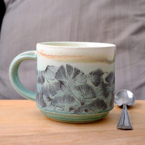 May include: A handmade ceramic mug with a light green handle and base. The mug features a botanical design of dark gray leaves against a light green background. The rim has a cream and tan color. A silver spoon rests on the wooden surface.