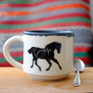 May include: A cream-colored ceramic mug with a dark gray rim and a black horse silhouette graphic. The mug features a subtle crackle glaze finish and a curved handle. A small silver-toned metal spoon rests beside the mug on a wooden surface.