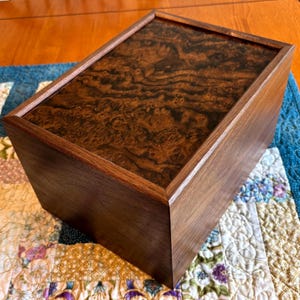 May include: A rectangular wooden box with a dark brown finish. The lid features a striking burl wood pattern, and the box is set on a colorful patchwork quilt. The box has a clean, modern design.