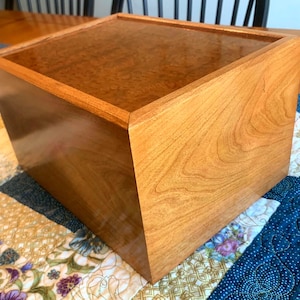 May include: A handcrafted wooden box with a glossy finish. The box is square with a flat top and is made of light brown wood. The wood grain is visible. The box is sitting on a floral and blue patterned table runner.