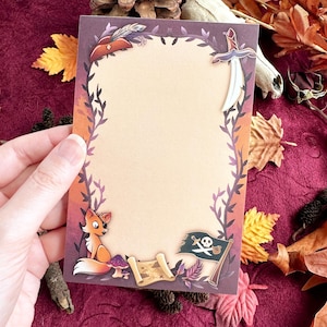 May include: A rectangular stationery card with a tan centre and a dark border featuring a fox wearing an eye patch, a pirate flag with a skull and crossbones, a treasure map, a sword, and a hat, all surrounded by dark purple vine illustrations.
