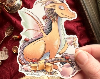 May include: A die-cut sticker featuring a light orange dragon with iridescent wings, holding a small teacup. The dragon is adorned with red roses and a long braided mane. Below are three brown paper bags, a teapot, and a teacup. The text reads @DreadPirateBri.
