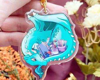 May include: A clear acrylic keychain featuring a teal dragon curled around a collection of art supplies, including a sketchbook, paintbrushes, and pencils. The dragon has white horns and scales, and the keychain includes a gold metal clasp.