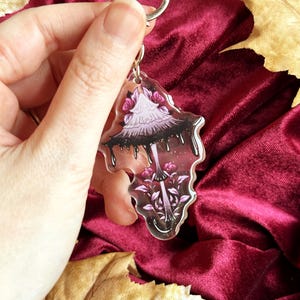 May include: A clear acrylic keychain with a mushroom design and a silver heart-shaped keyring. The mushroom has a light purple cap with black dripping details, pink roses, and a pink stem. The background is a deep red velvet.