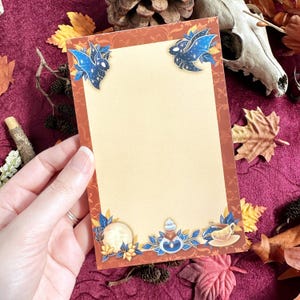 May include: A rectangular notepad with a tan center and a dark orange border featuring a floral pattern. The corners and bottom edge are decorated with illustrations of blue bats, a full moon, a potion bottle, and a teacup with blue leaves.