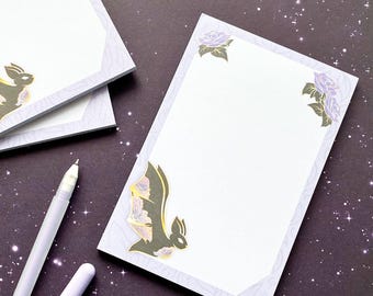 Op de afbeelding: Een rechthoekig notitieblok met een wit midden en een lichtpaarse rand met een vleermuisillustratie in de linkerbenedenhoek en paarse roosillustraties in de bovenhoeken. Een witte pen zonder dop ligt naast het notitieblok op de ondergrond.