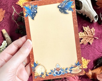 Op de afbeelding: Een rechthoekig notitieblok met een beige centrum en een donkeroranje rand met een decoratief patroon. De hoeken en de onderrand zijn versierd met illustraties van blauwe sterrenvleermuizen, een maan, een flesje en een theekopje met blaadjes.