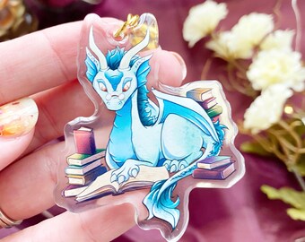 May include: A clear acrylic charm featuring a light blue dragon with white horns and small glasses, depicted reading an open book while surrounded by several stacks of colorful books. The charm includes a gold-toned metal jump ring at the top.