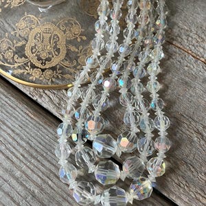 May include: A multi-strand necklace featuring clear, faceted crystal beads with an iridescent finish. The beads are arranged in several cascading lengths, reflecting light with rainbow hues. The necklace is composed of round, cut glass beads strung together, creating a shimmering, reflective appearance throughout the entire piece of jewelry.
