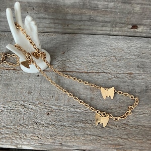 May include: A gold-colored necklace with three tooth-shaped pendants. The necklace is displayed on a white ceramic hand against a weathered wooden background. The chain is delicate and the teeth are detailed.