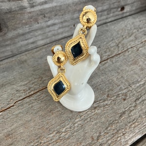 May include: Gold-tone clip-on earrings with a geometric design. Each earring features a round top and a diamond-shaped pendant with a dark blue center and a rope-like border. The earrings are displayed on a white ceramic hand.