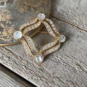 May include: A gold-toned brooch with a geometric design. The brooch features a central diamond shape with four large, clear, round stones at each corner. The design is embellished with smaller, clear stones. The brooch is set against a wooden surface.