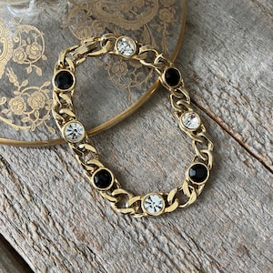 May include: A gold-tone chain link bracelet featuring alternating round black stones and clear faceted crystals set within the metal links. The bracelet has a secure clasp mechanism and a polished finish, showcasing a repeating pattern of dark and light accents.