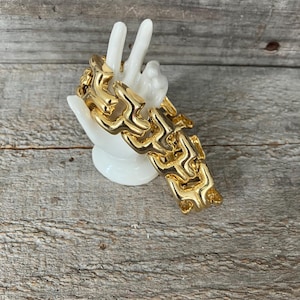 May include: A gold-toned metal bracelet featuring a series of interlocking, stylized geometric links with a polished, reflective finish, displayed on a white ceramic hand-shaped stand.