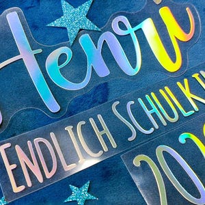 Könnte beinhalten: Holografische Vinyl-Aufkleber mit dem Namen Henri, dem Text ENDLICH SCHULKIND! und der Jahreszahl 2026. Die Schriftzüge haben eine reflektierende, mehrfarbige Oberfläche, die auf transparentem Untergrund zwischen Blau, Lila und Gelb schimmert.