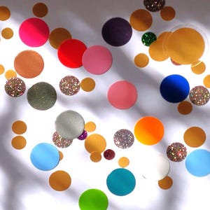 Könnte beinhalten: Eine Ansammlung farbenfroher, runder Dekorationselemente, die auf einer weißen Oberfläche verstreut sind. Die Elemente umfassen verschiedene Größen und Farben, wie Gold, Rosa, Blau und glitzernde Texturen, wodurch eine verspielte und künstlerische Anordnung entsteht.