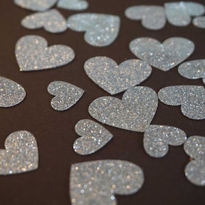 Könnte beinhalten: Eine Ansammlung von silbernen, glitzernden Herz-Ausschnitten auf einer dunkelbraunen Oberfläche. Die Herzen sind unterschiedlich groß und haben eine glitzernde, strukturierte Oberfläche. Ideal für Bastelarbeiten und Dekorationen.