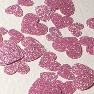 Könnte beinhalten: Eine Ansammlung von pinkfarbenen, glitzernden Herz-Ausschnitten auf einer weißen Oberfläche. Die Herzen, in verschiedenen Größen, sind mit funkelndem Glitzer bedeckt. Ideal für Bastelarbeiten oder dekorative Zwecke.
